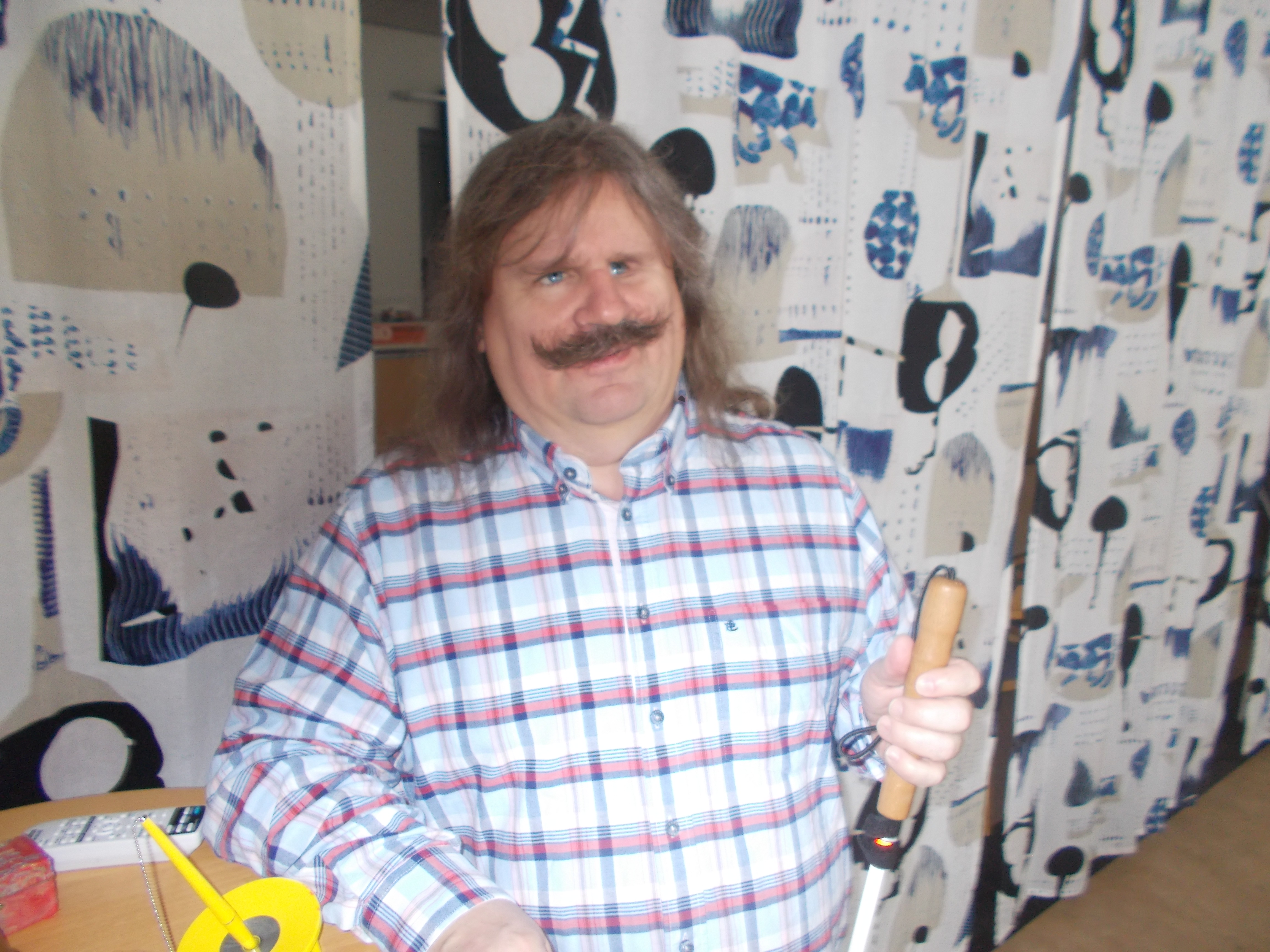 Thomsson har långt gråsprängt hår och en kraftig mustach. Blå,röd,vitrutig skjorta och håller den vita käppen i vänster hand