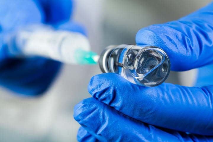 Bild på vaccinations dos i flaska och en spruta som blå handsk klädda händer håller i