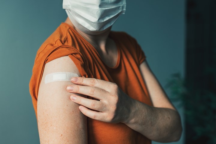En kvinna visar upp sin högra överarm med ett plåster på efter vaccination. Hon är munskydd och har en rostfärgad topp.