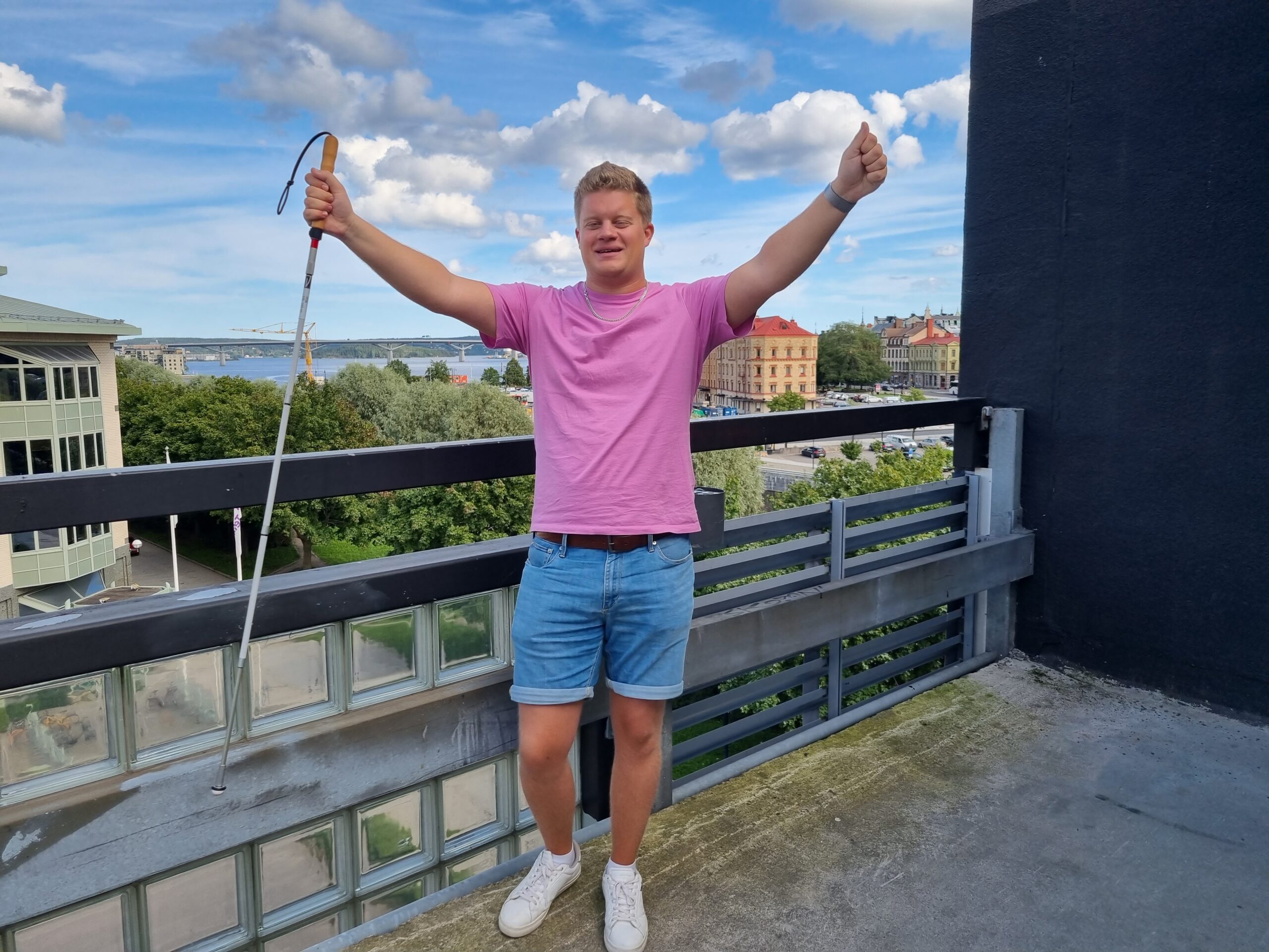 Mattias gör segergest med vita käppen i ena handen. Han bär en rosa t-shirt, blå jeansshorts och vita snikers. I bakgrunden Sundsvallsbron och Kulturmagasinet oc