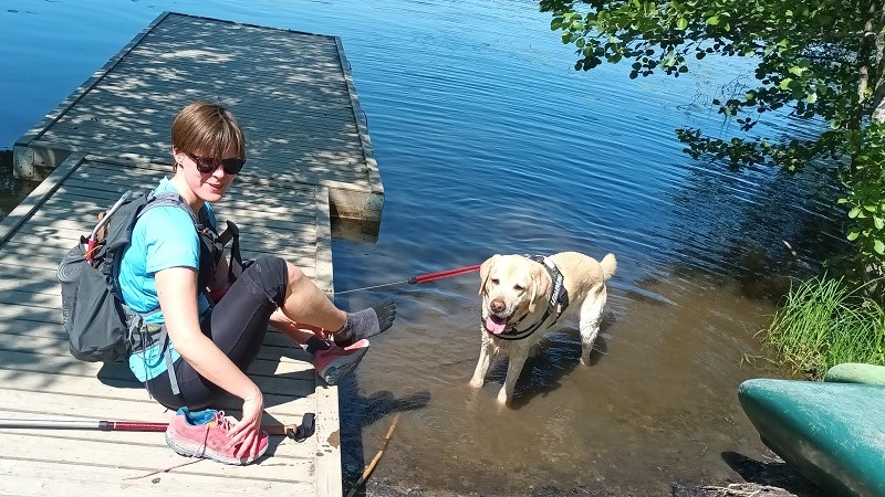 Ida har brunt kortklippt hår och mörka glasögon. Hon sitter på en träbrygga vid en sjö men ryggsäcken på ryggen. I vattnet nedanför står hennes golden retriver som är hennes ledarhund.